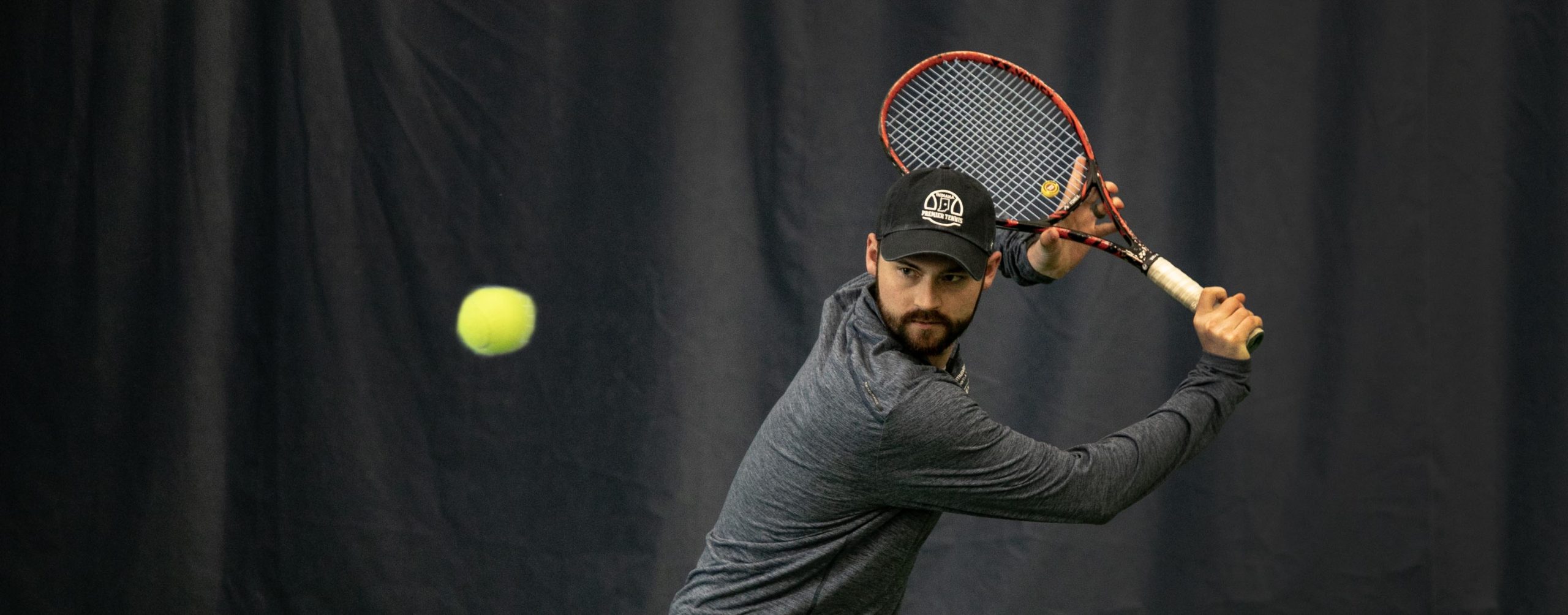Male in black hat and grey long sleeve shirt focuses on hitting the tennis ball.