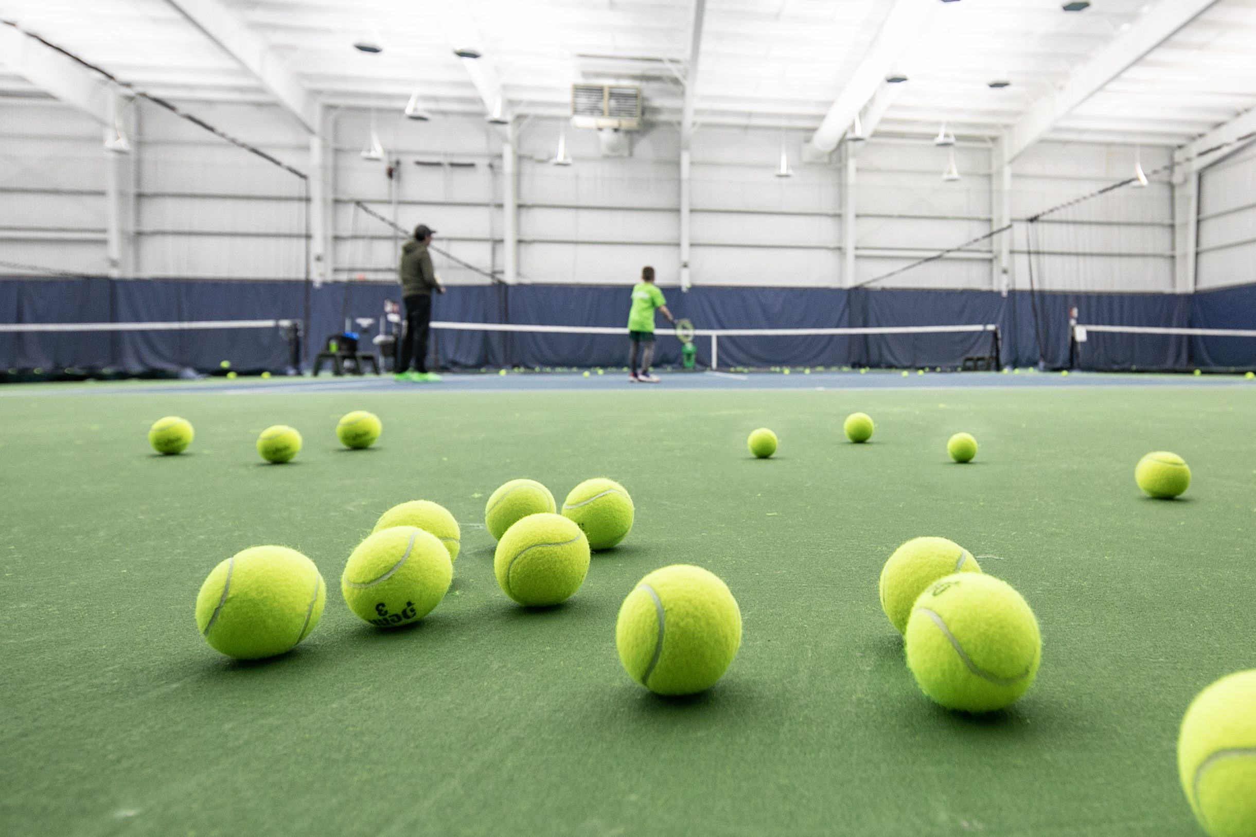 Tennis balls laying across the tennis court with two tennis players in the background.